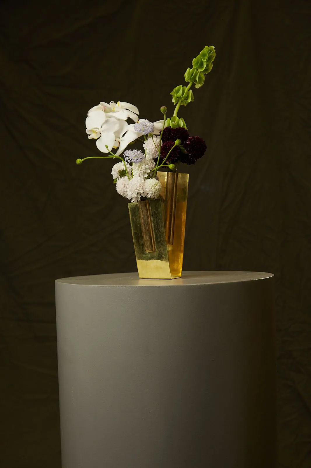 Vaso-escultura Vértice do Ouro em latão, com flores brancas, sobre pedestal cinza