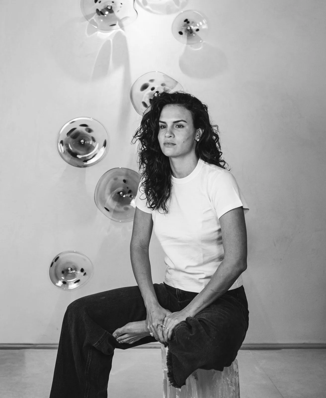 Retrato de Marcela Helena sentada diante de peças de vidro na parede